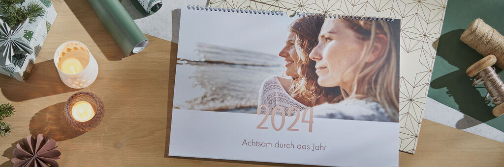 Auf einem Tisch liegt ein Fotokalender im Querformat. Das Titelblatt zeigt zwei Frauengesichter und im Hintergrund das Meer. Darunter steht „2022: Achtsam durch das Jahr“. Neben dem Kalender sind Teelichter, Weihnachtsdekoration und passende Geschenkverpackungsaccessoires zu sehen.